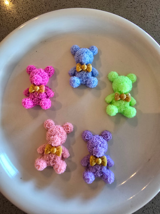 Cinco adorables ositos decorativos hechos completamente de pequeñas rosas de espuma, dispuestos en un plato blanco. Los ositos están en colores pastel: rosa fucsia vibrante, celeste suave, verde menta, rosa pálido y lila. Cada osito tiene un elegante moño dorado en el cuello. Las rosas que forman cada figura están densamente agrupadas creando una textura uniforme y suave. Los ositos están fotografiados desde arriba sobre una superficie clara, mostrando la calidad artesanal y los detalles de cada pieza. 