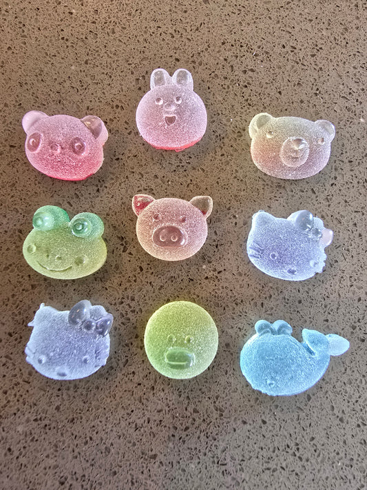 Set de 9 charms kawaii holográficos translúcidos dispuestos en círculo: osito rosa, conejito blanco, osito beige, ranita verde, cerdito rosa, Hello Kitty azul, Hello Kitty morada, círculo verde y ballena azul con efectos iridiscentes sobre superficie beige 