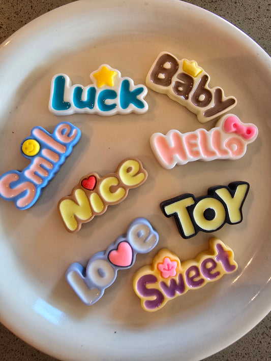 Set de figuras decorativas con palabras en inglés como "Luck", "Baby", "Smile", "Hello", "Nice", "Love", "TOY" y "Sweet" en colores vibrantes sobre plato blanco, perfectas para decoden y manualidades kawaii con mensajes positivos


