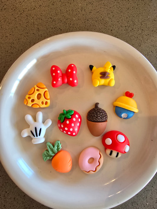 Set de 10 charms temáticos incluyendo queso suizo amarillo, Pikachu, lazo rojo con lunares, fresa roja, hongo de Mario Bros, dona rosa, zanahoria naranja, bellota marrón y guante de Mickey Mouse en plato blanco, perfectos para decoden kawaii y manualidades gaming 