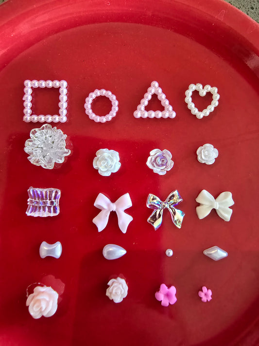 Set completo de 20 piezas decorativas para nail art en tonos rosa y nácar sobre fondo rojo, mostrando formas geométricas con perlas, flores de diferentes tamaños, lazos elegantes, perlas sueltas y pequeñas flores, organizadas en filas para mostrar la variedad del conjunto

