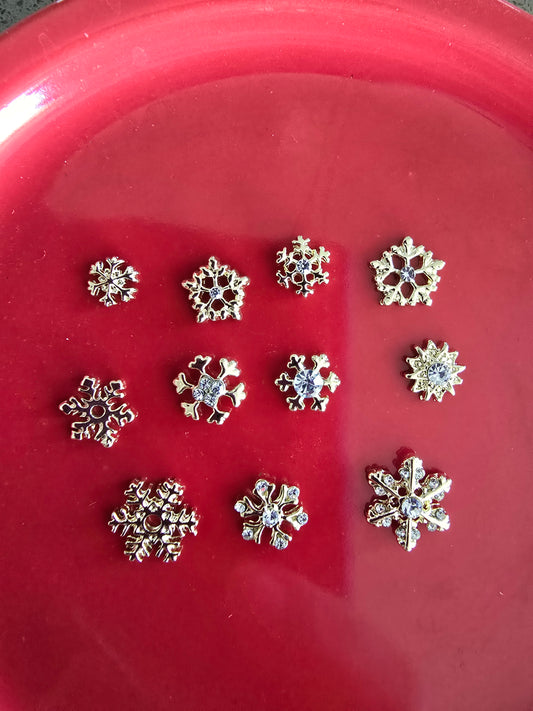 Set de 10 copos de nieve plateados con cristales centrales brillantes en bandeja roja, mostrando diferentes patrones geométricos únicos con acabados metálicos, perfectos para nail art invernal y decoración navideña premium 