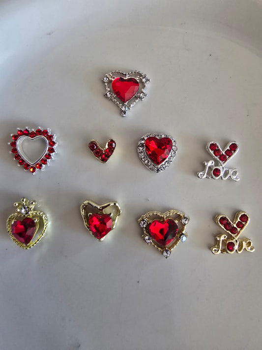 Set de 10 corazones decorativos rojos con cristales para nail art sobre fondo blanco, mostrando diferentes diseños incluyendo corazones con strass dorados y plateados, cristales rojos centrales y dijes con la palabra Love

