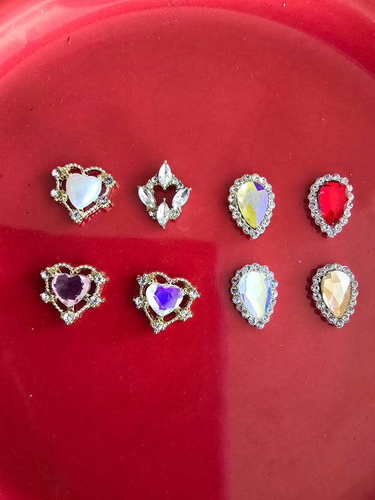Set de 10 gemas decorativas con formas de corazón y lágrima en colores vibrantes como azul, morado, rojo y amarillo con engastes dorados sobre fondo rojo, perfectas para nail art premium y manualidades de lujo 